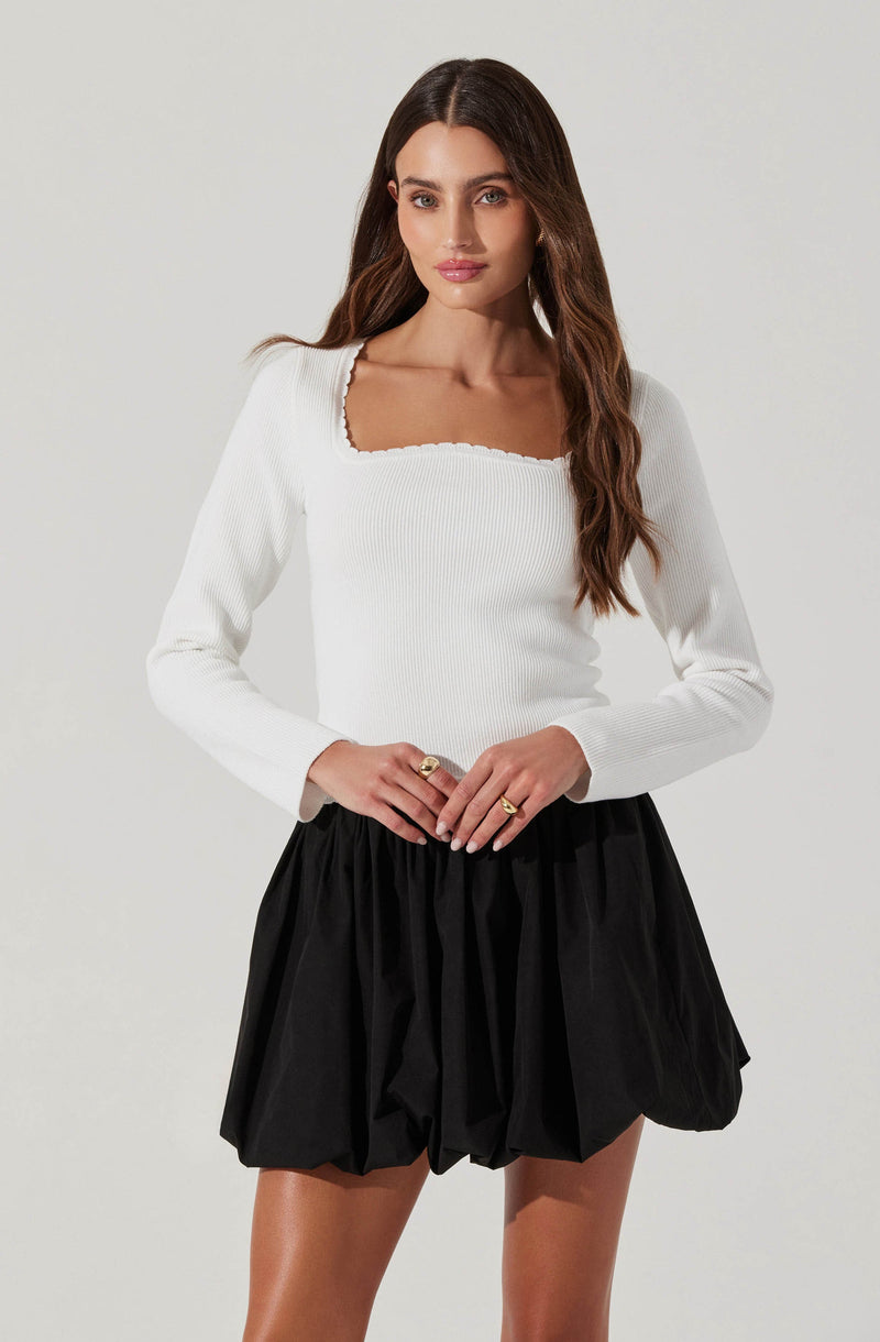 A woman is wearing a long-sleeved, white, ribbed top with a square neckline, paired with a black, voluminous, pleated mini skirt.