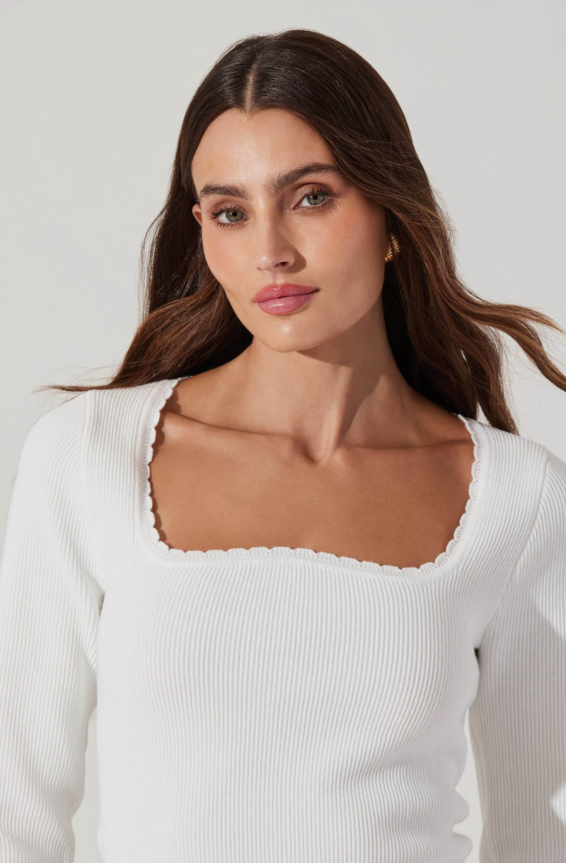 A woman is wearing a white, long-sleeved, ribbed top with a square neckline that features a ruffled edge detail.