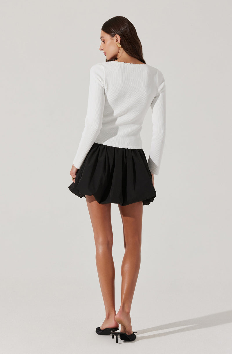 A woman is dressed in a long-sleeved white top with a slightly textured fabric and a round neckline, paired with a black, voluminous, gathered mini skirt that has a bubbled, cloud-like appearance. She is also wearing black pointed-toe heels with small bows on the front.