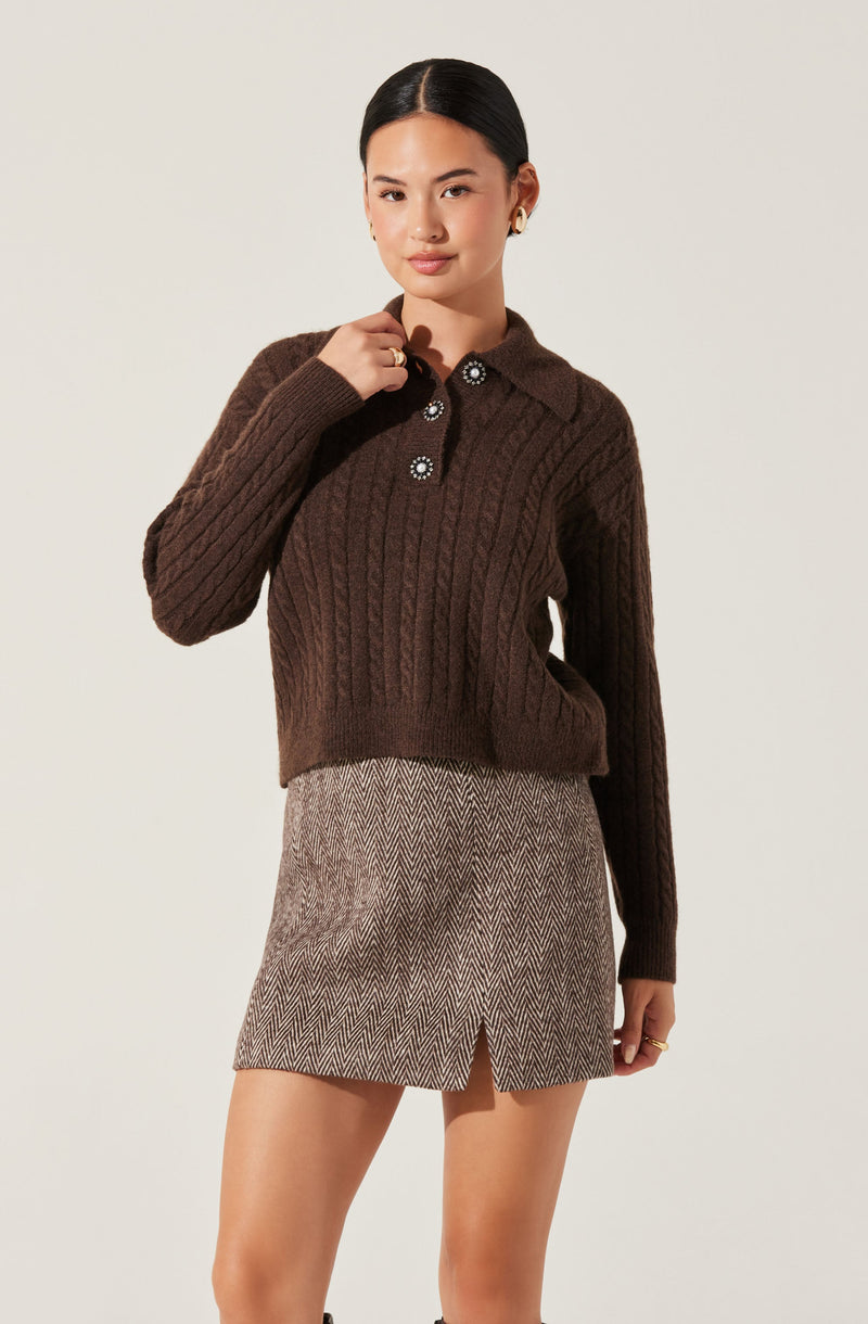 A woman is wearing a dark brown, chunky knit sweater with a cable pattern. The sweater features a collared neckline with four decorative buttons, each adorned with a flower-like design, and long sleeves. She also wears a beige and brown herringbone patterned skirt with a small slit in the front.
