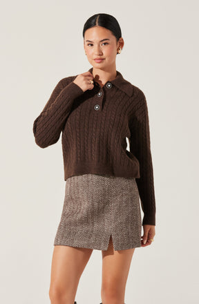 A woman is wearing a dark brown, chunky knit sweater with a cable pattern. The sweater features a collared neckline with four decorative buttons, each adorned with a flower-like design, and long sleeves. She also wears a beige and brown herringbone patterned skirt with a small slit in the front.