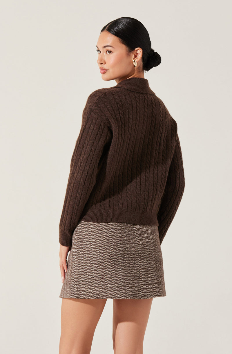 A woman is wearing a dark brown, cable-knit sweater with a high neckline and long sleeves. She pairs it with a high-waisted, mini skirt in a beige and brown herringbone pattern. She has her dark hair styled in a low bun and is accessorized with gold hoop earrings.