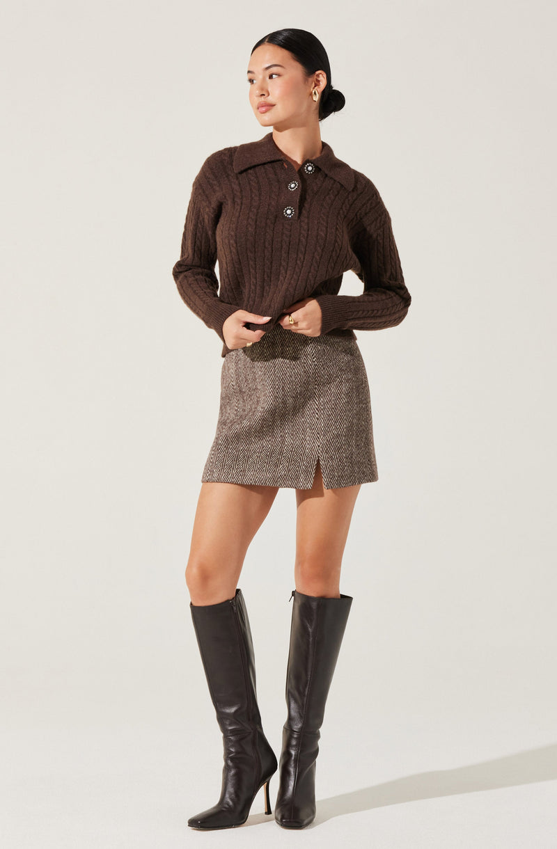 A woman is wearing a dark brown, long-sleeved, cable-knit sweater with a collar and four buttons on the front. She pairs it with a high-waisted, herringbone-patterned skirt in shades of gray that has a front slit. She completes the look with knee-high black leather boots with stiletto heels.