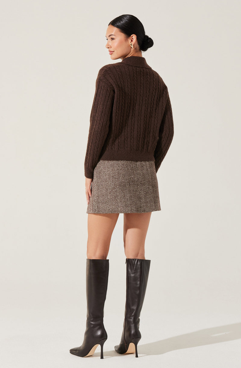 A woman is wearing a dark brown, cable-knit sweater with a high collar and long sleeves. She pairs it with a fitted, herringbone-patterned mini skirt in neutral tones. She completes her outfit with knee-high, black leather stiletto boots.