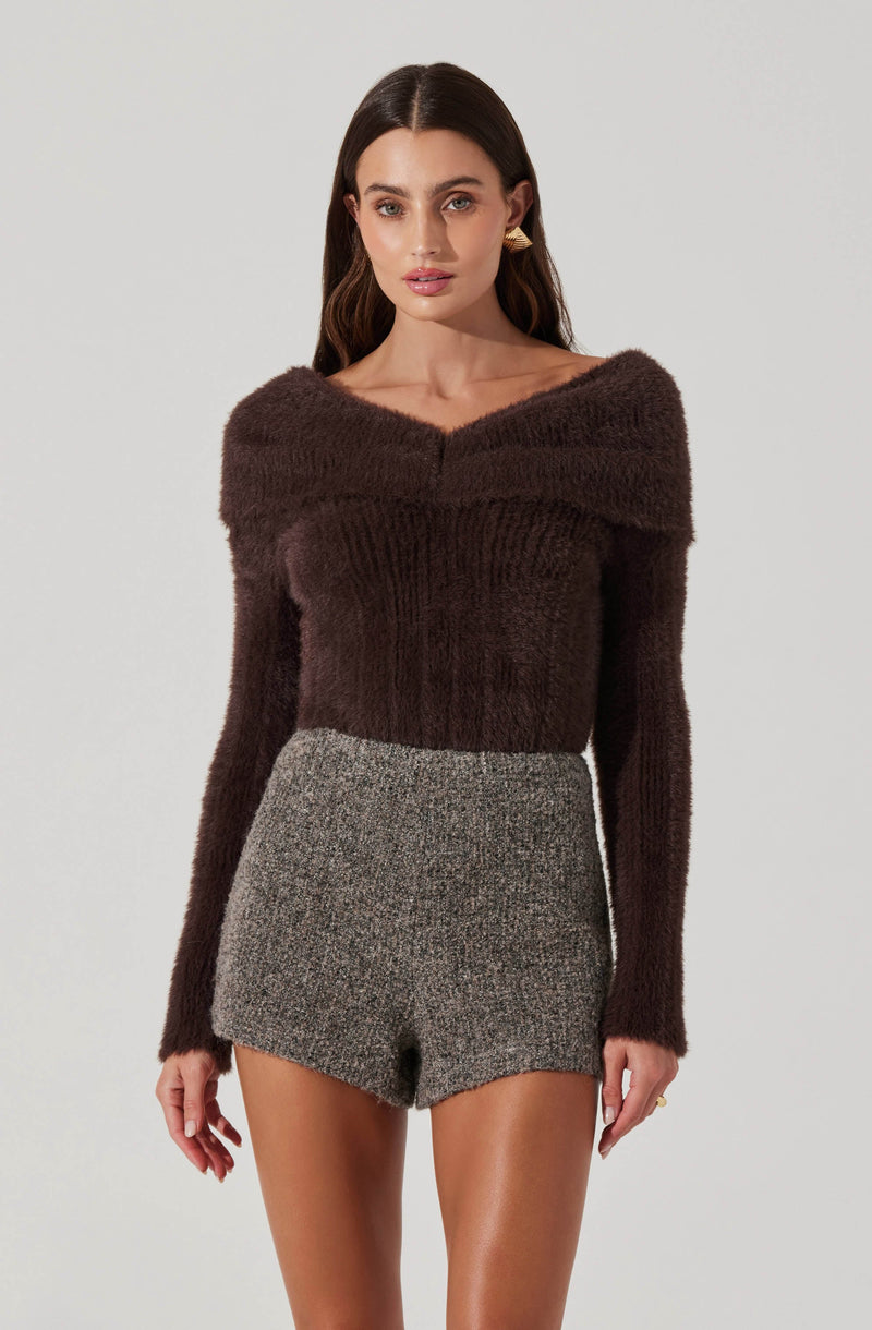 A woman is wearing a dark brown, long-sleeved, fuzzy sweater with a wide neckline that drapes off the shoulders. She is also wearing high-waisted, gray, textured shorts.