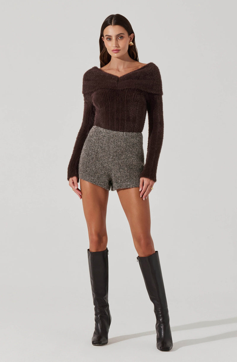 A woman is wearing a brown, off-the-shoulder, long-sleeve sweater that appears to be made of a fuzzy, textured material. She pairs it with high-waisted, gray, tweed shorts. Her outfit is completed with knee-high black leather boots.