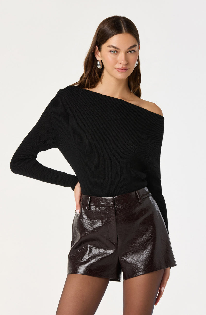 A woman is wearing a black, off-the-shoulder, long-sleeve knit top paired with high-waisted, shiny black leather shorts. She accessorizes with large, patterned earrings.