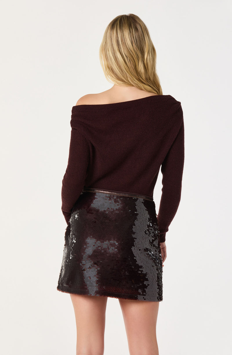 A woman is wearing an off-the-shoulder, long-sleeve, burgundy knit sweater paired with a high-waisted, black sequined skirt.