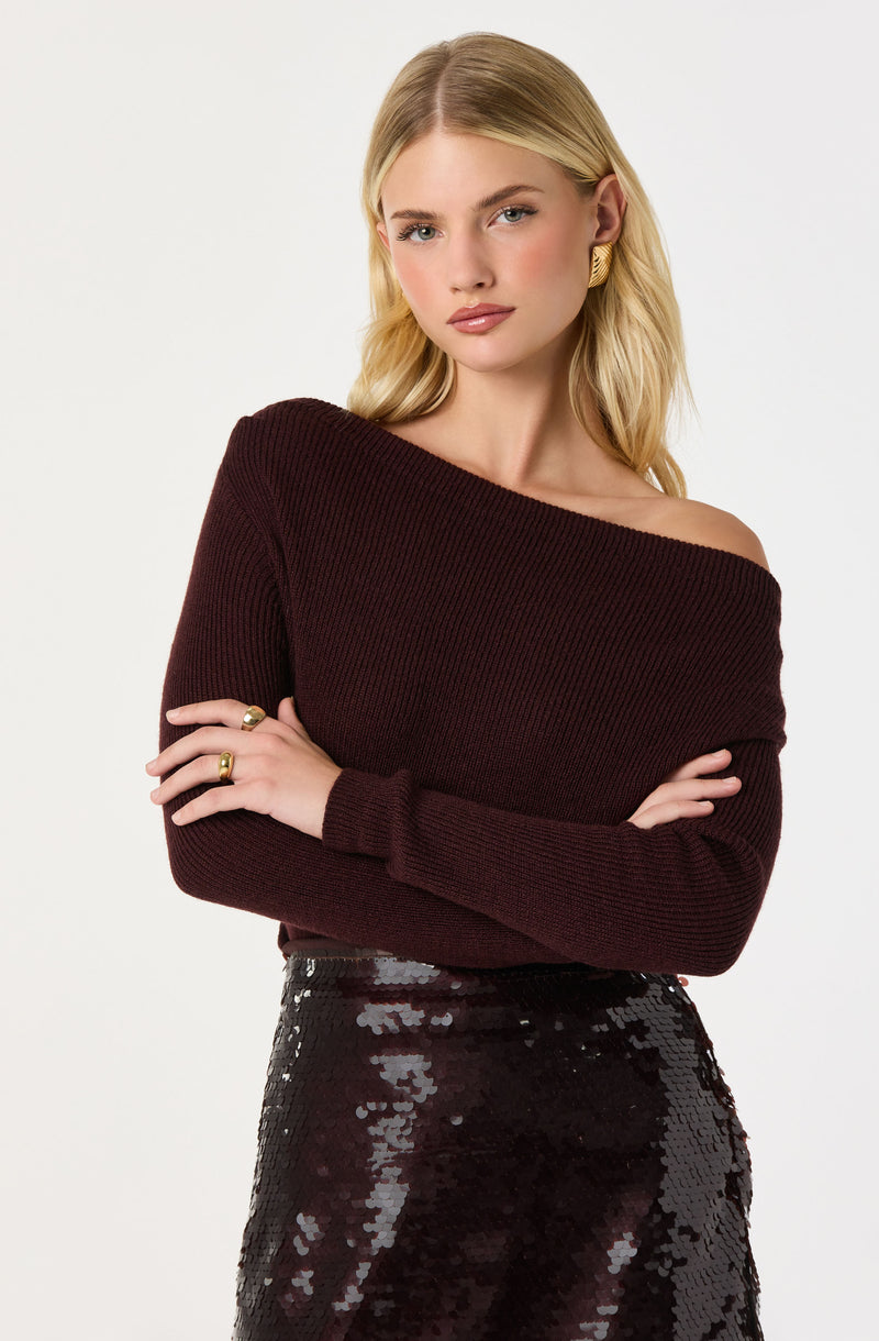 A young woman with blonde hair is wearing an off-the-shoulder, long-sleeved, dark burgundy knitted sweater paired with a high-waisted, black sequin skirt. She has gold earrings and rings on her fingers.