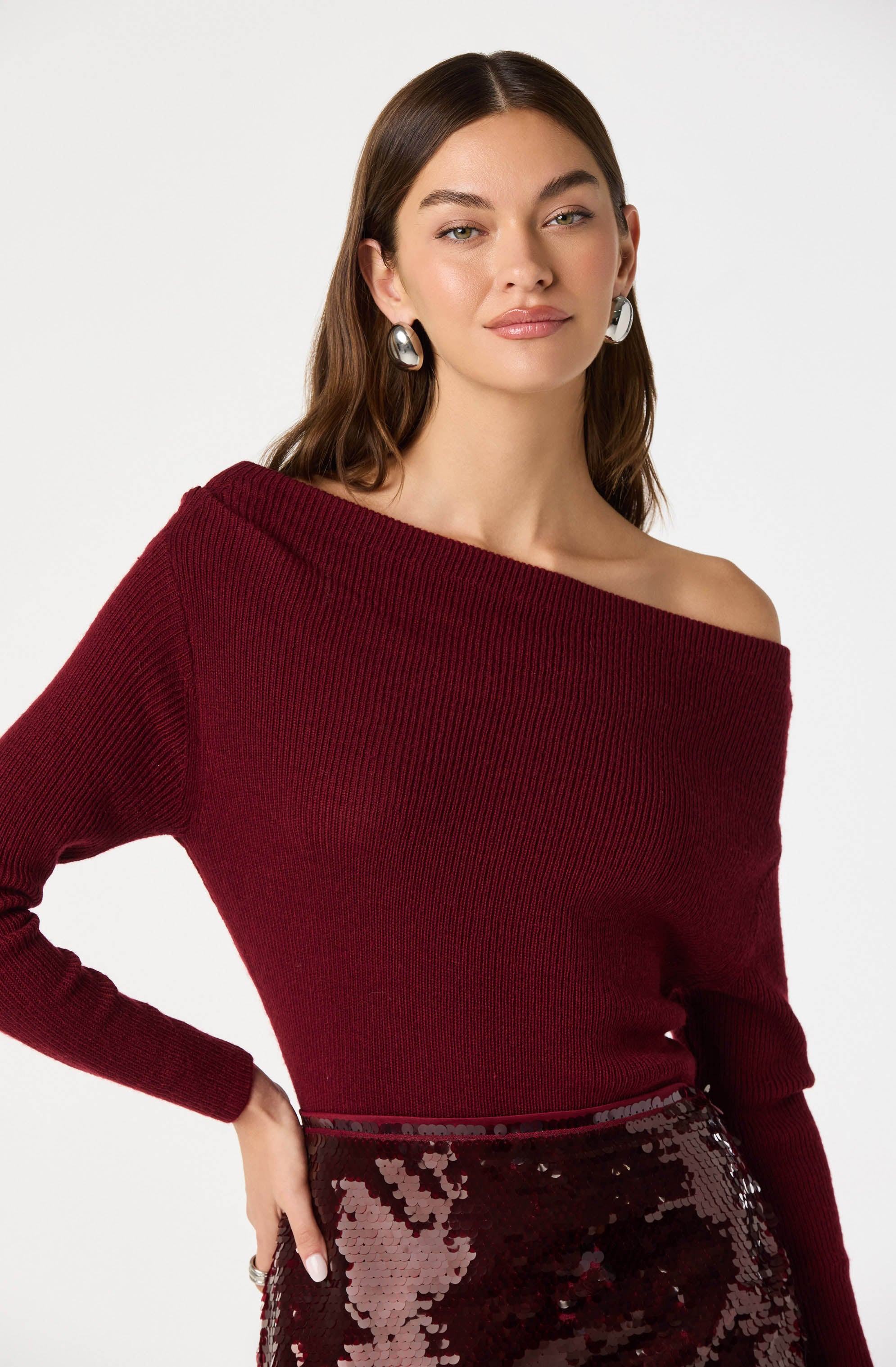 A woman is wearing an off-the-shoulder, long-sleeved, deep red knit sweater and a high-waisted, sequined skirt with a pattern of darker and lighter red sequins. She is accessorized with large, oval-shaped silver earrings.