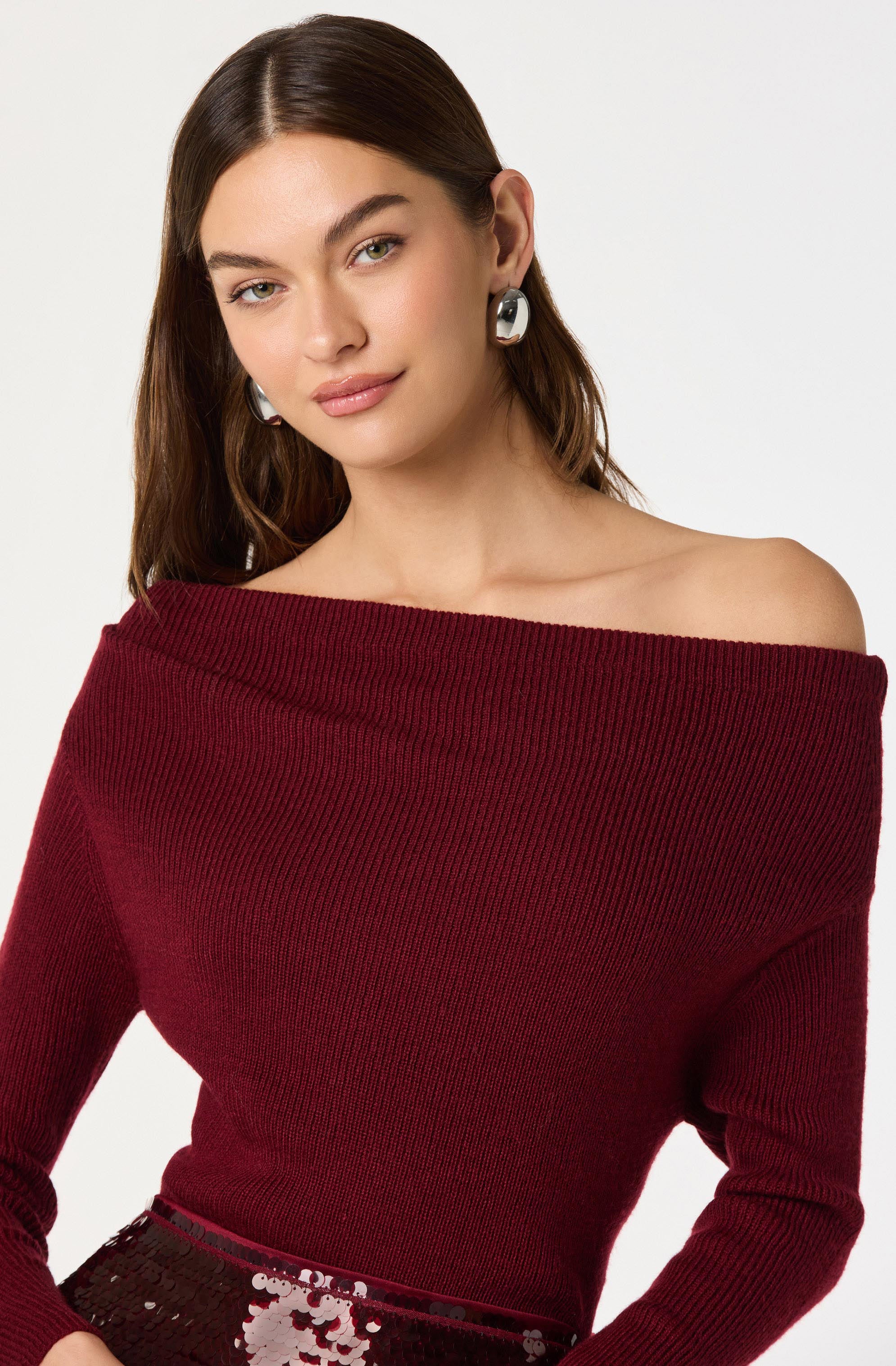A woman is wearing an off-the-shoulder, long-sleeved, ribbed sweater in a deep red color. She has paired it with large, geometric silver earrings and a skirt with sequins.