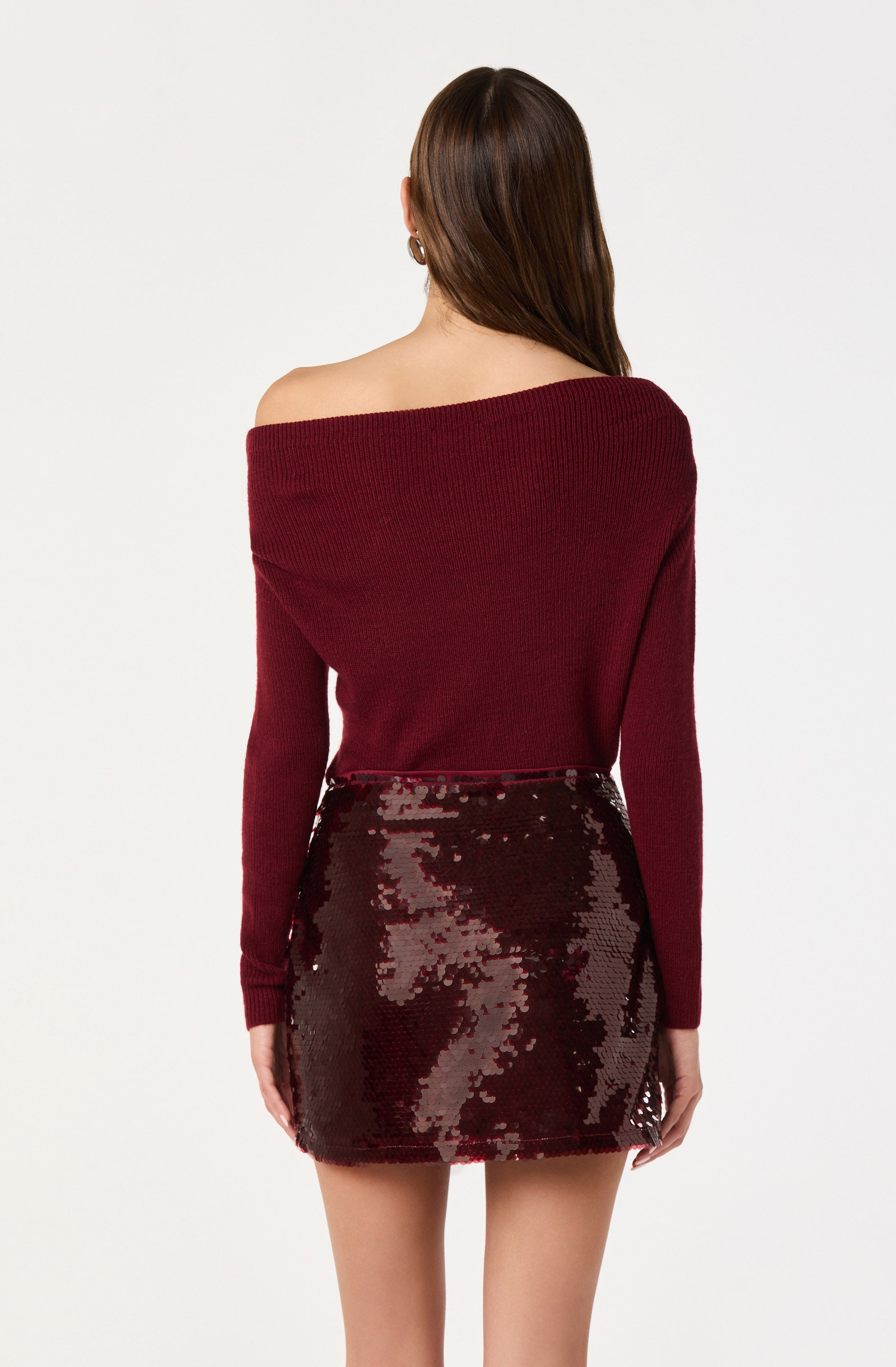 A person is wearing an off-the-shoulder, long-sleeve, dark red sweater paired with a high-waisted, shiny, sequined mini skirt in a similar dark red color.