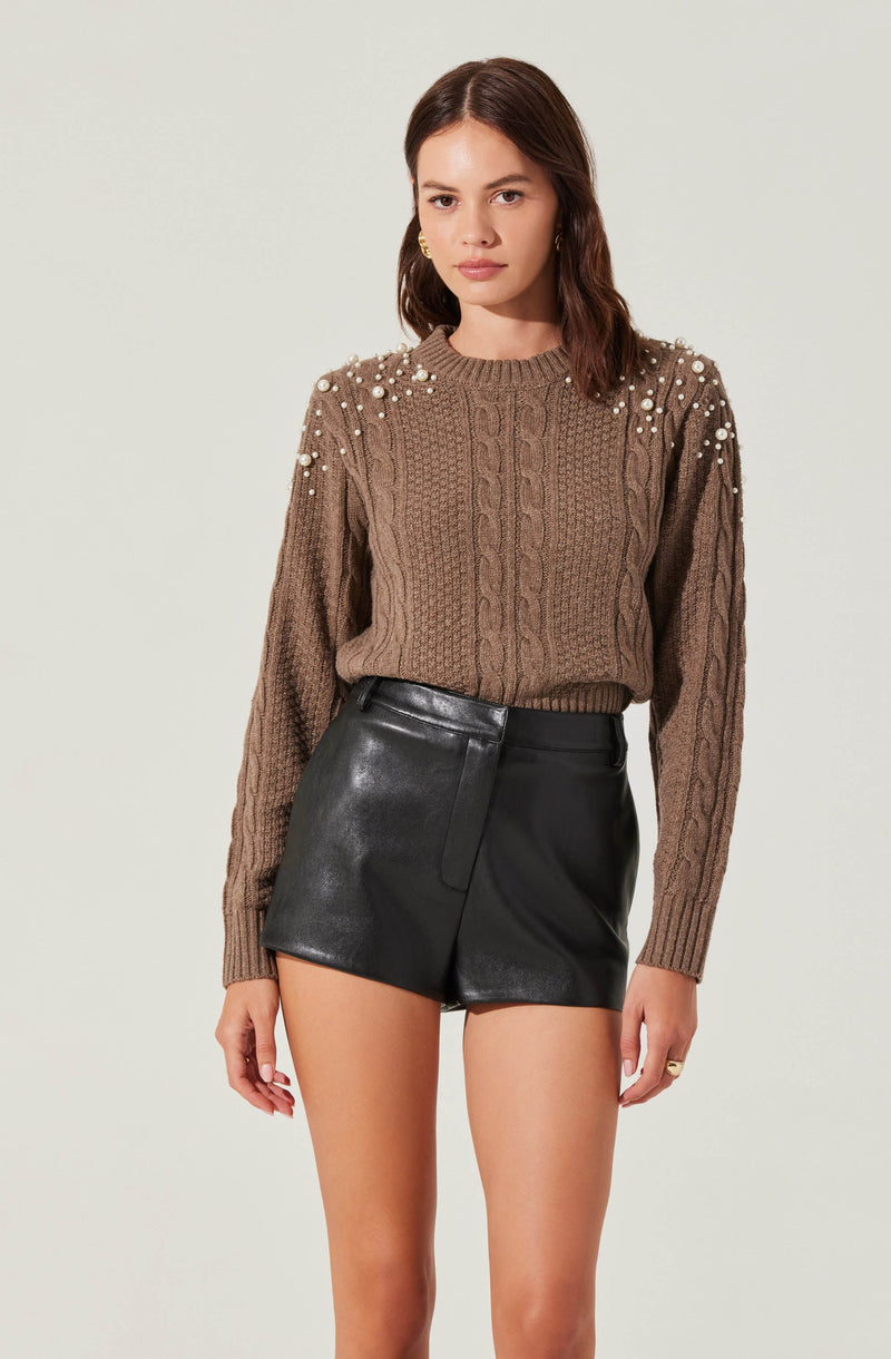 A woman is wearing a brown cable-knit sweater embellished with small white pearls on the shoulders and upper sleeves. She pairs the sweater with high-waisted black leather shorts.