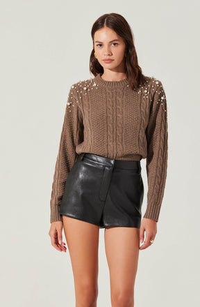 A woman is wearing a brown cable-knit sweater embellished with small white pearls on the shoulders and upper sleeves. She pairs the sweater with high-waisted black leather shorts.