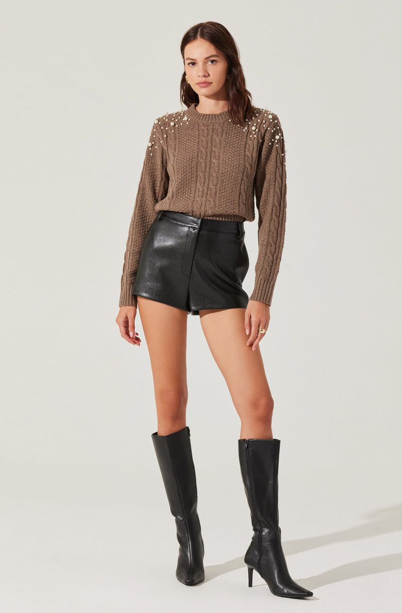 A woman is wearing a brown cable-knit sweater with pearl embellishments on the shoulders, paired with high-waisted black leather shorts. She is also wearing tall black leather heeled boots and gold hoop earrings.