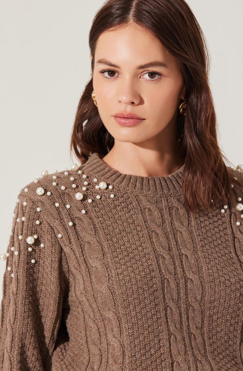 A woman wearing a brown knitted sweater featuring a cable knit pattern. The sweater has pearl embellishments on the shoulders, adding decorative detail. She is also wearing gold earrings.