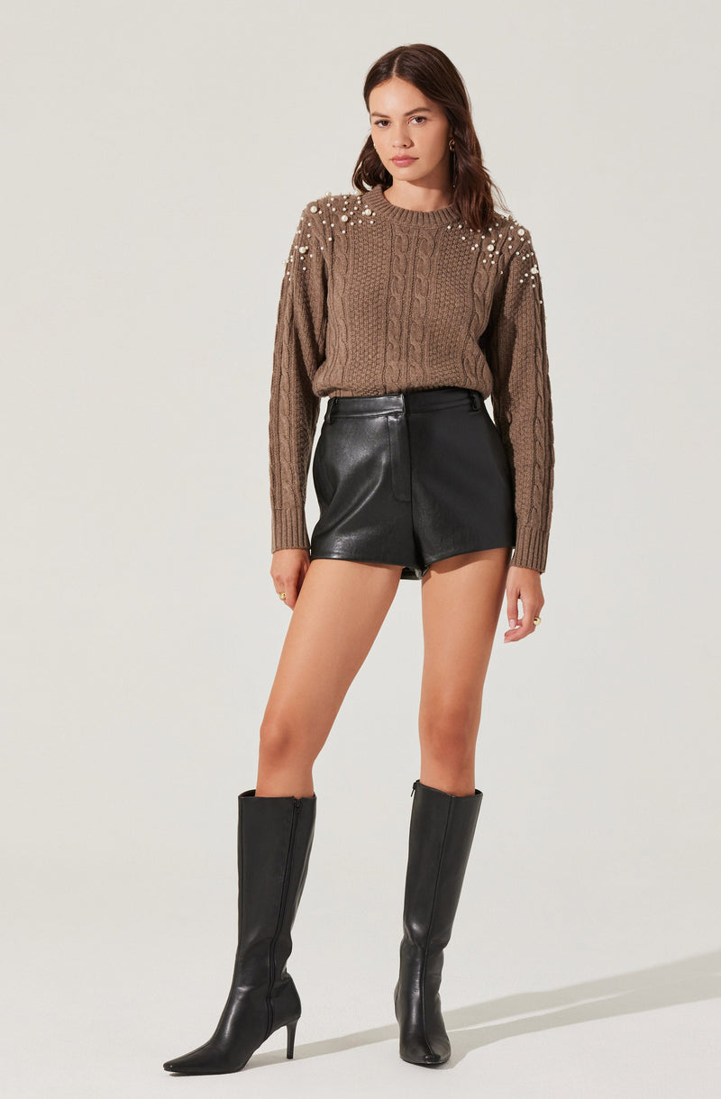 A woman is wearing a brown knit sweater with pearl embellishments on the shoulders and upper arms. She pairs it with high-waisted black leather shorts and black knee-high heeled boots.