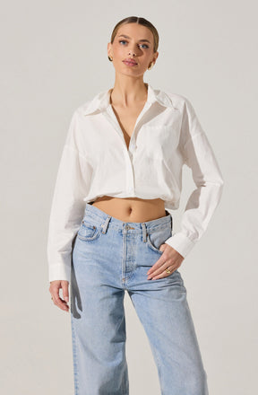 A woman is wearing a white, long-sleeved, button-down shirt that is slightly oversized and tied at the waist, paired with light blue, high-waisted jeans. The shirt has a classic collar and a front pocket on the left side.