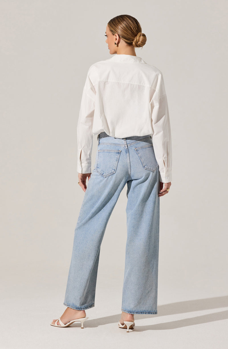 A woman is wearing a white long-sleeve button-up shirt with a loose fit, tucked into light-wash high-waisted jeans with a relaxed, wide-leg cut. She is also wearing white heeled sandals.