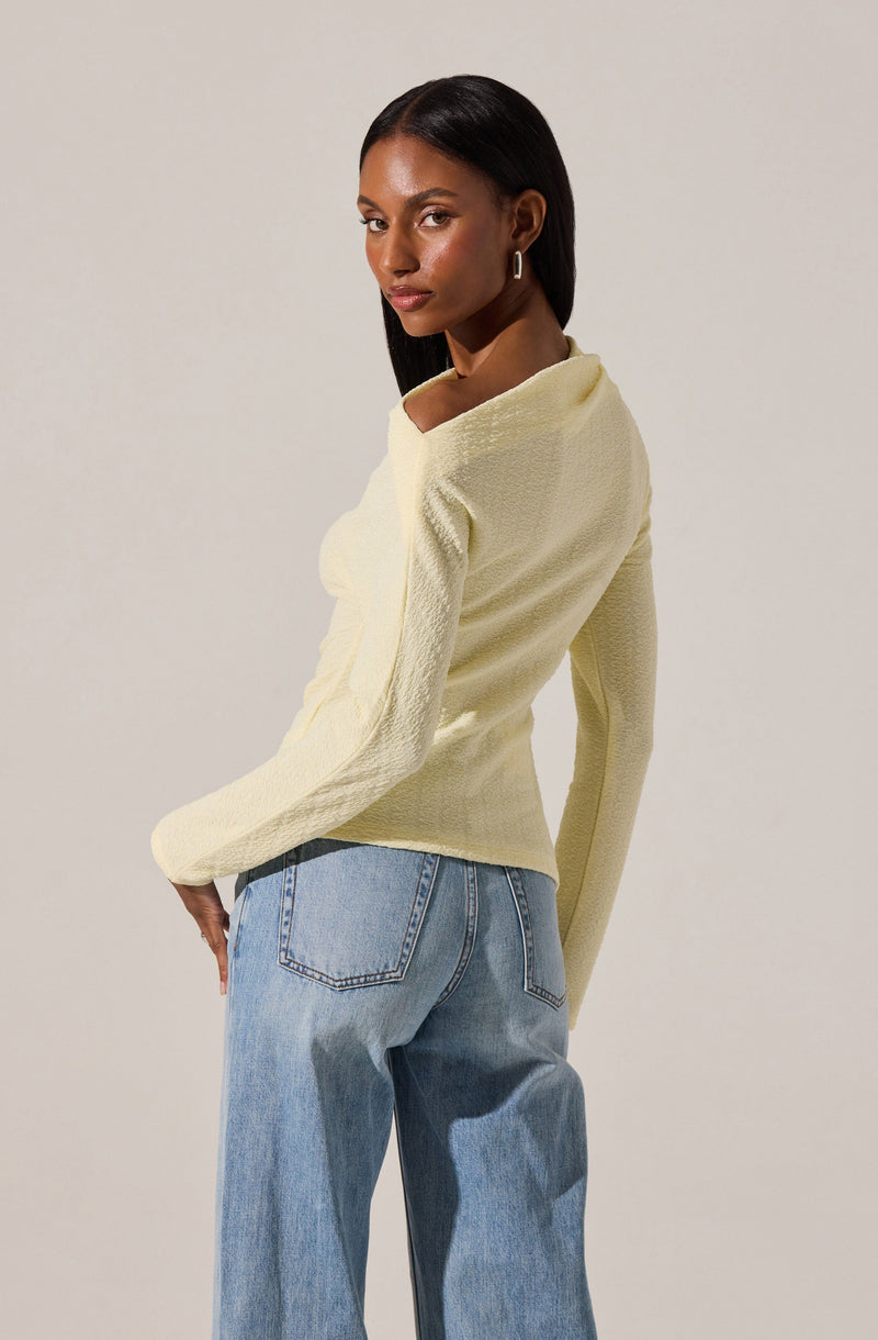A woman with dark skin and straight, shoulder-length black hair is wearing a light yellow textured long-sleeve top with a wide neckline, paired with high-waisted, loose-fitting light blue jeans. She is also accessorized with small hoop earrings.