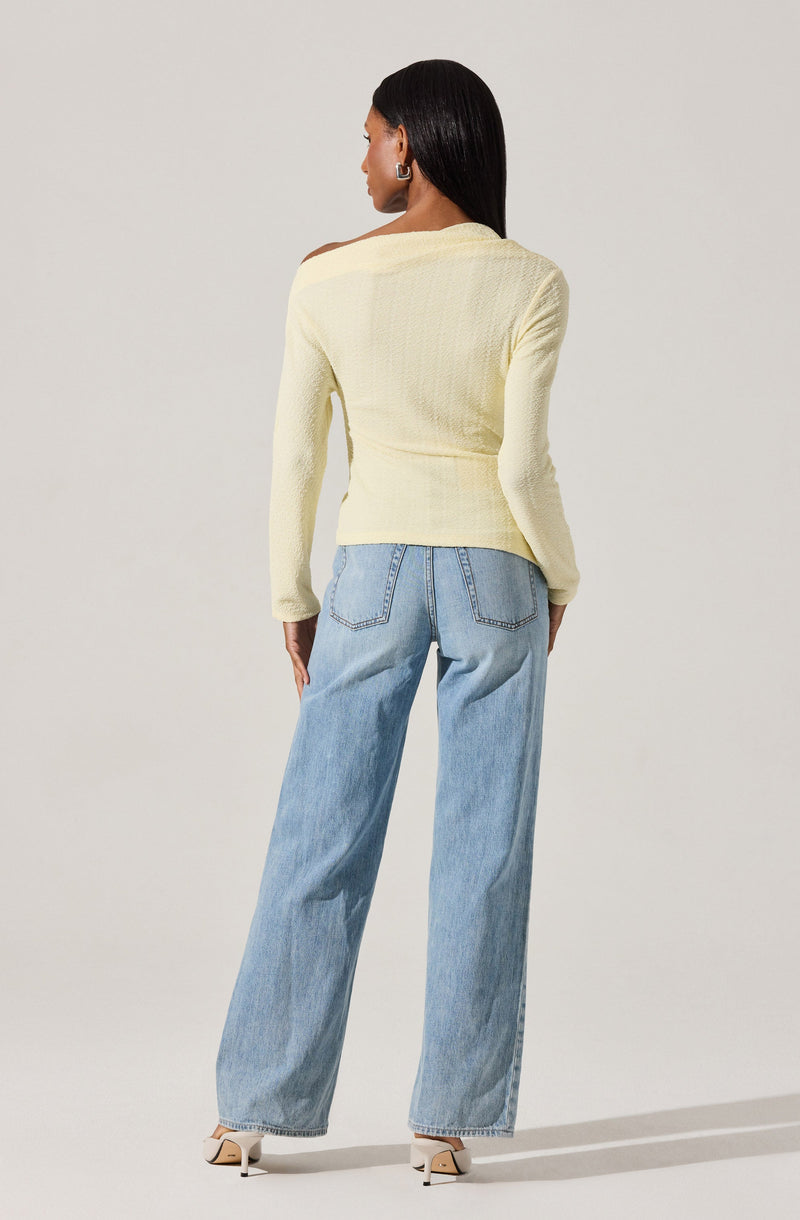 A woman is wearing a light yellow textured knit sweater with long sleeves and a wide neckline that extends over one shoulder. She pairs the sweater with high-waisted, light-wash, wide-leg jeans that have back pockets. She completes the outfit with beige high-heeled shoes.