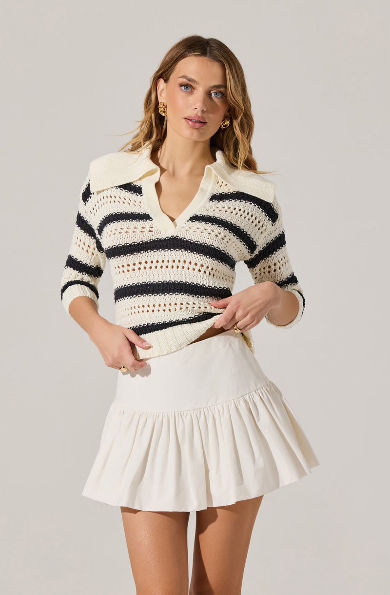 A woman is wearing a cream-colored, knit sweater with black horizontal stripes and a wide collar. She is also wearing a cream-colored, high-waisted skirt with a ruffled hem.