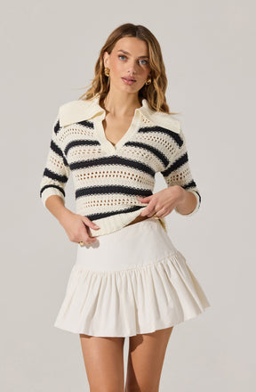 A woman is wearing a cream-colored, knit sweater with black horizontal stripes and a wide collar. She is also wearing a cream-colored, high-waisted skirt with a ruffled hem.