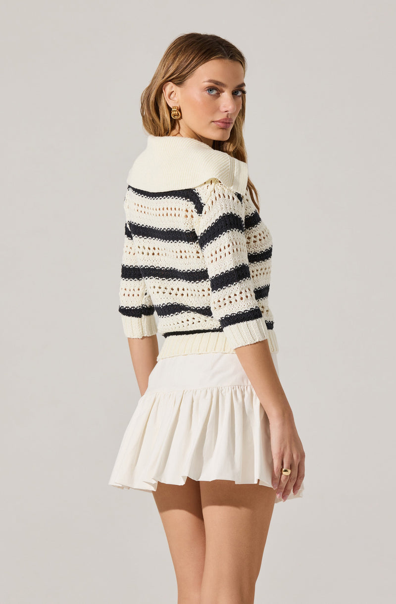 A woman is wearing a cream-colored knit sweater with horizontal navy blue and orange stripes and an oversized collar. She pairs it with a white, pleated, ruffled skirt.