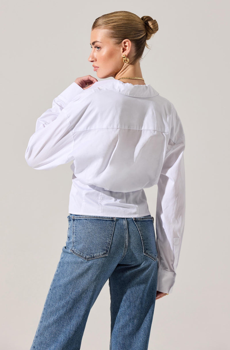 A woman is wearing a long-sleeved white button-up shirt with a collar. The shirt is slightly oversized, with a loose fit around the waist, and the sleeves are gathered at the cuffs. She pairs the shirt with high-waisted blue jeans that have a classic five-pocket design.