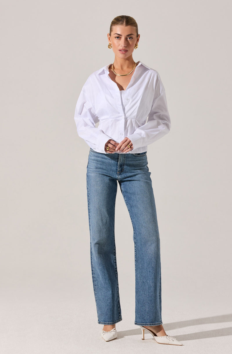 A woman is wearing a white button-up shirt with long sleeves and a gathered detail at the waist. She pairs the shirt with high-waisted, straight-leg blue jeans. She is accessorized with gold hoop earrings, layered gold necklaces, and rings. She is wearing pointed white heels.