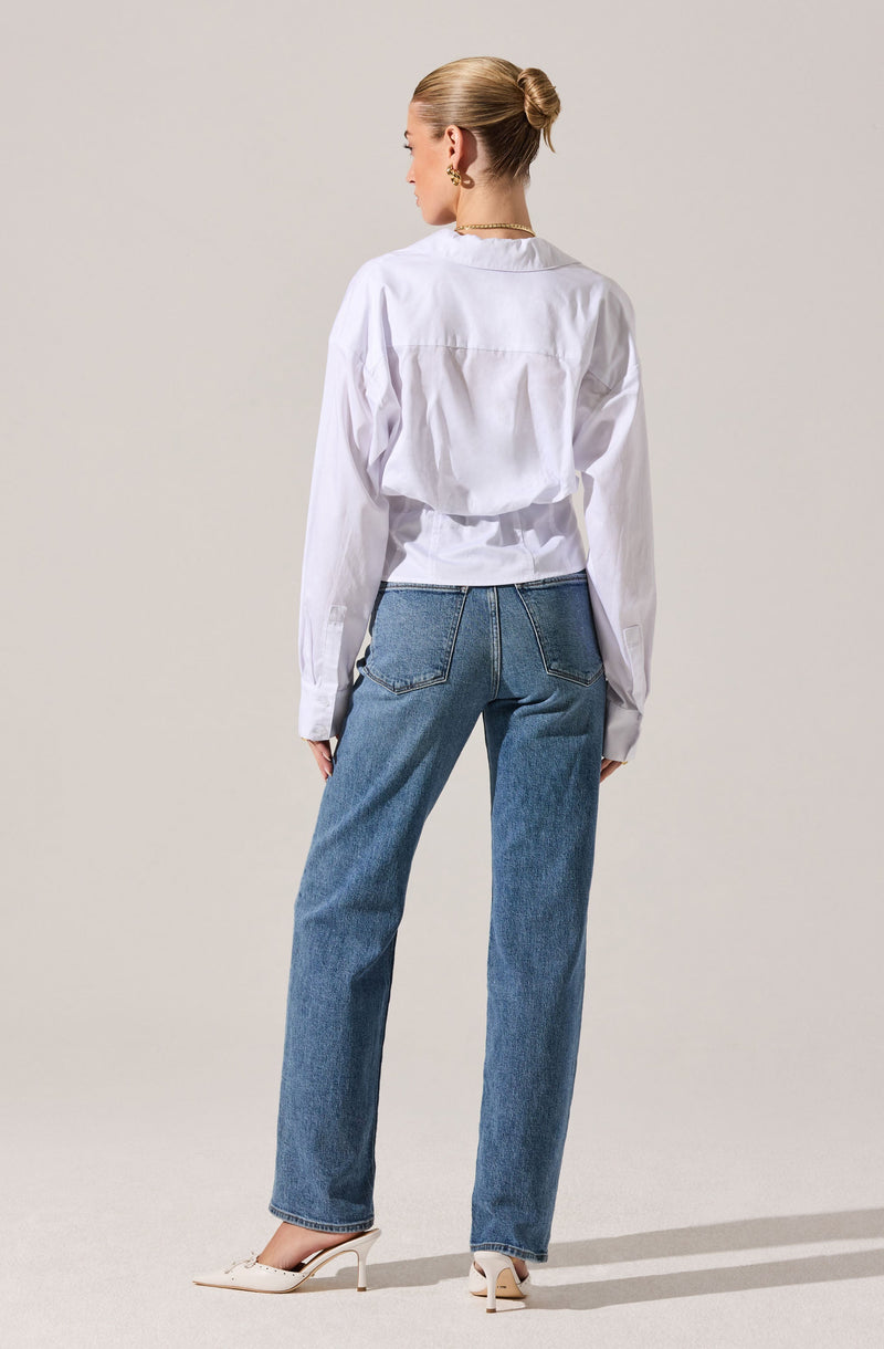 A person is wearing a loose-fitting, long-sleeved white shirt with a slightly oversized fit and a collared neckline. They are also wearing high-waisted, straight-leg blue jeans with visible stitching on the back pockets. The outfit is completed with a pair of beige high-heeled shoes. The person has their hair styled in a neat bun and is accessorized with earrings and a necklace.