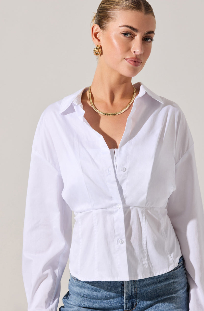 A woman is wearing a white button-up blouse with a gathered waistline and a collar. The blouse has long sleeves, and she has left the top few buttons undone. She is accessorized with gold hoop earrings and a gold chain necklace. She is also wearing blue jeans.