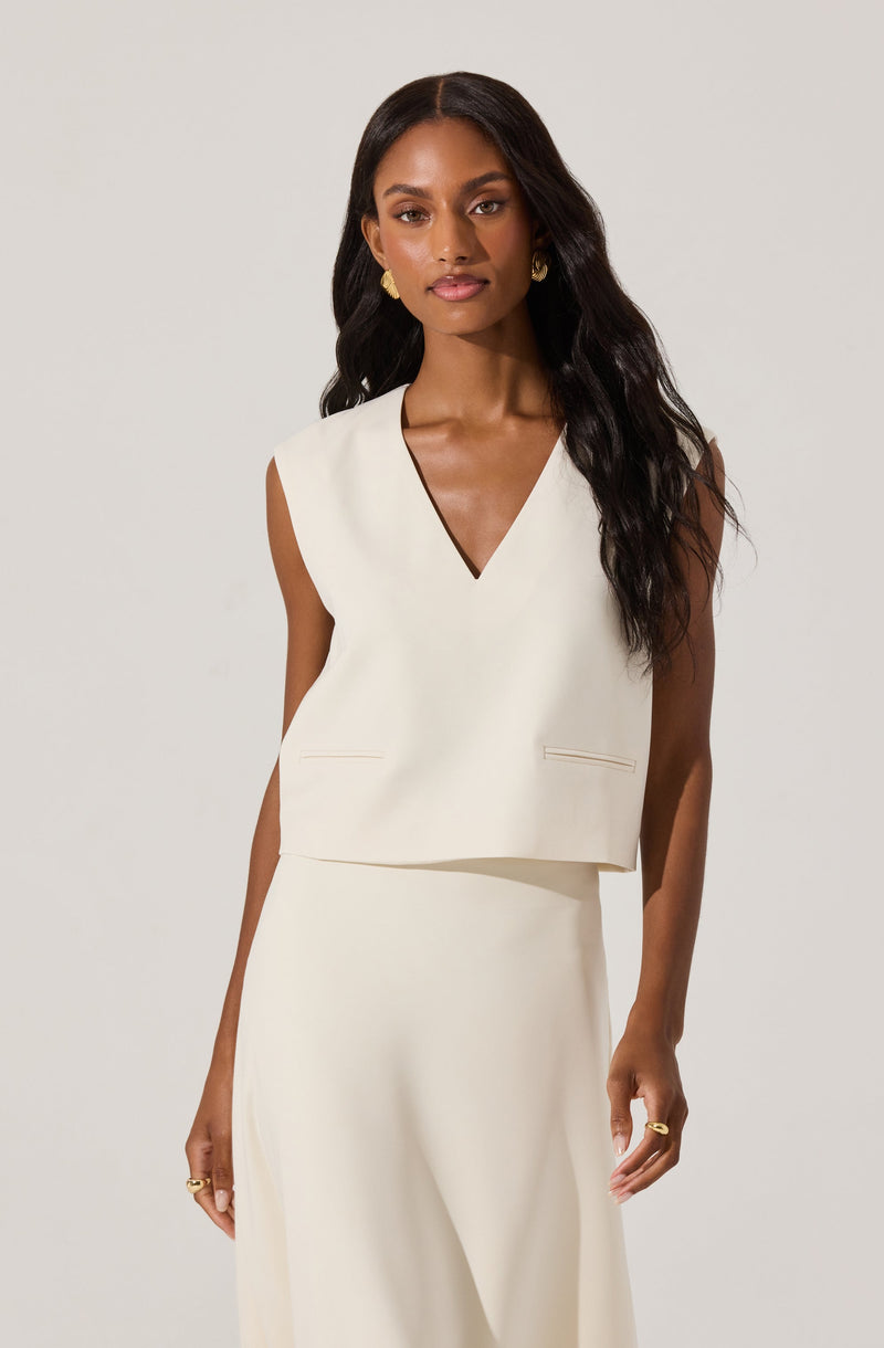 A woman is wearing a sleeveless, cream-colored outfit consisting of a V-neck top with a boxy fit and two small front pockets, paired with a matching high-waisted skirt. She accessorizes with gold earrings and rings.