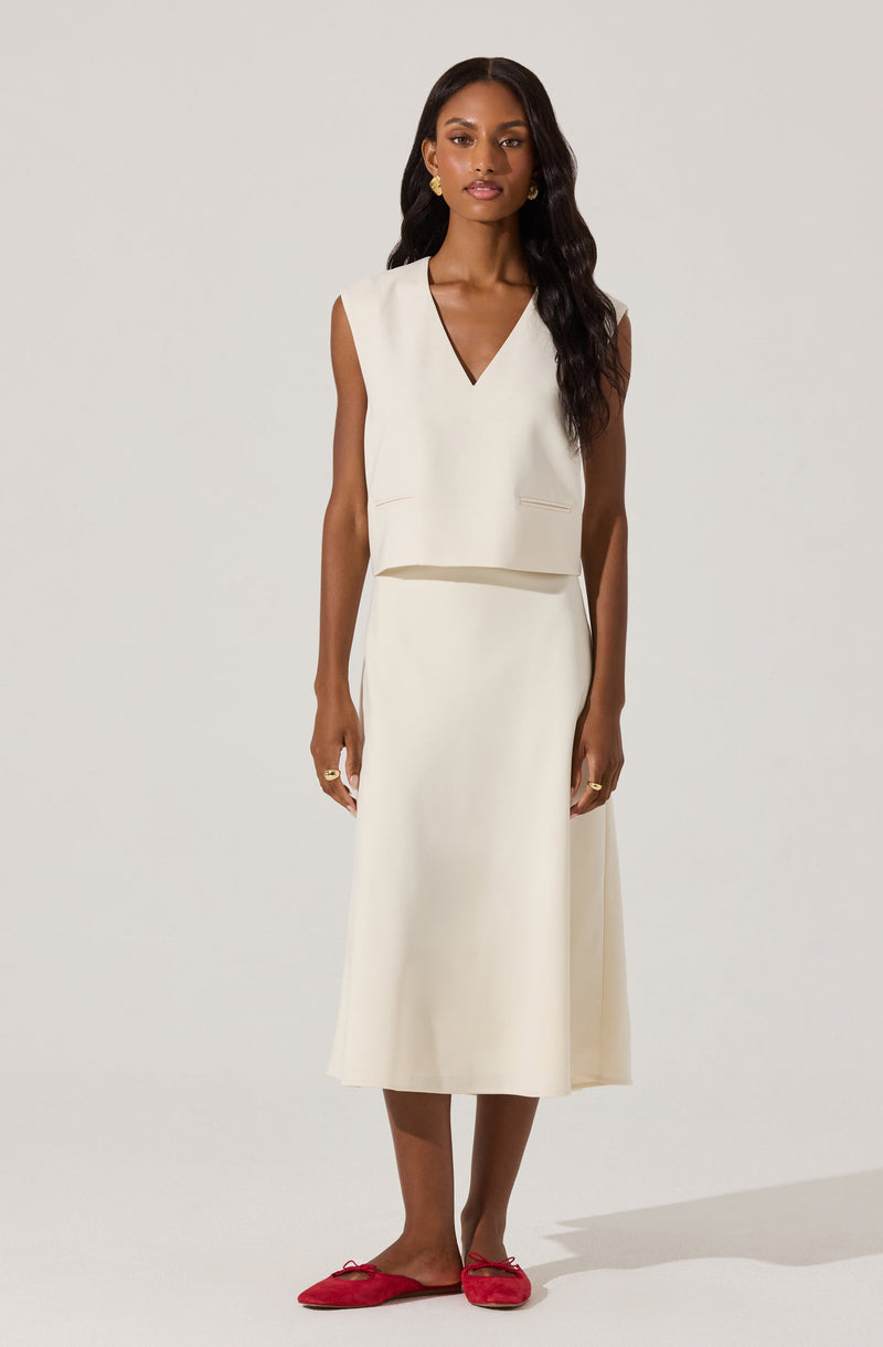 The woman is wearing a sleeveless white top with a V-neckline and matching high-waisted white skirt that falls below the knees. She has on gold earrings and rings, and is barefoot with red slippers.