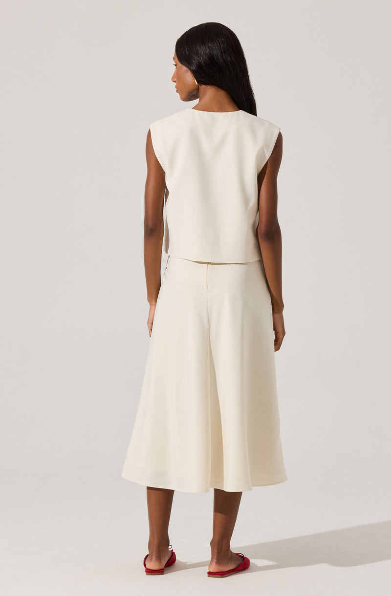 A woman is wearing a sleeveless, off-white, cropped top with wide shoulder details, paired with a matching high-waisted, wide-leg skirt that falls below the knees. She is also wearing red flat sandals.