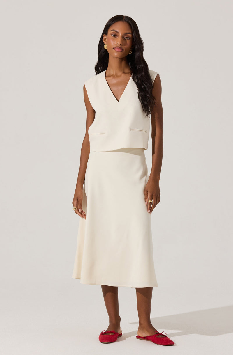 A woman is wearing a sleeveless, cream-colored top with a V-neckline and subtle lapel details at the front. She pairs it with a matching cream-colored, midi-length skirt that has a simple, clean silhouette. She completes the outfit with red flats featuring a bow detail on the top. She also accessorizes with gold stud earrings and rings on both hands.