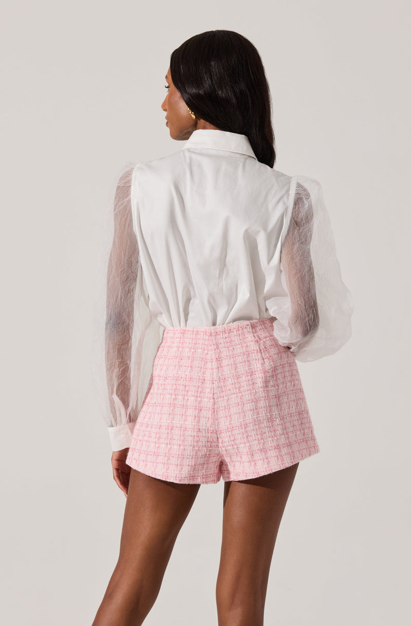A woman is wearing a white button-up shirt with sheer, puffed long sleeves, and high-waisted pink tweed shorts.