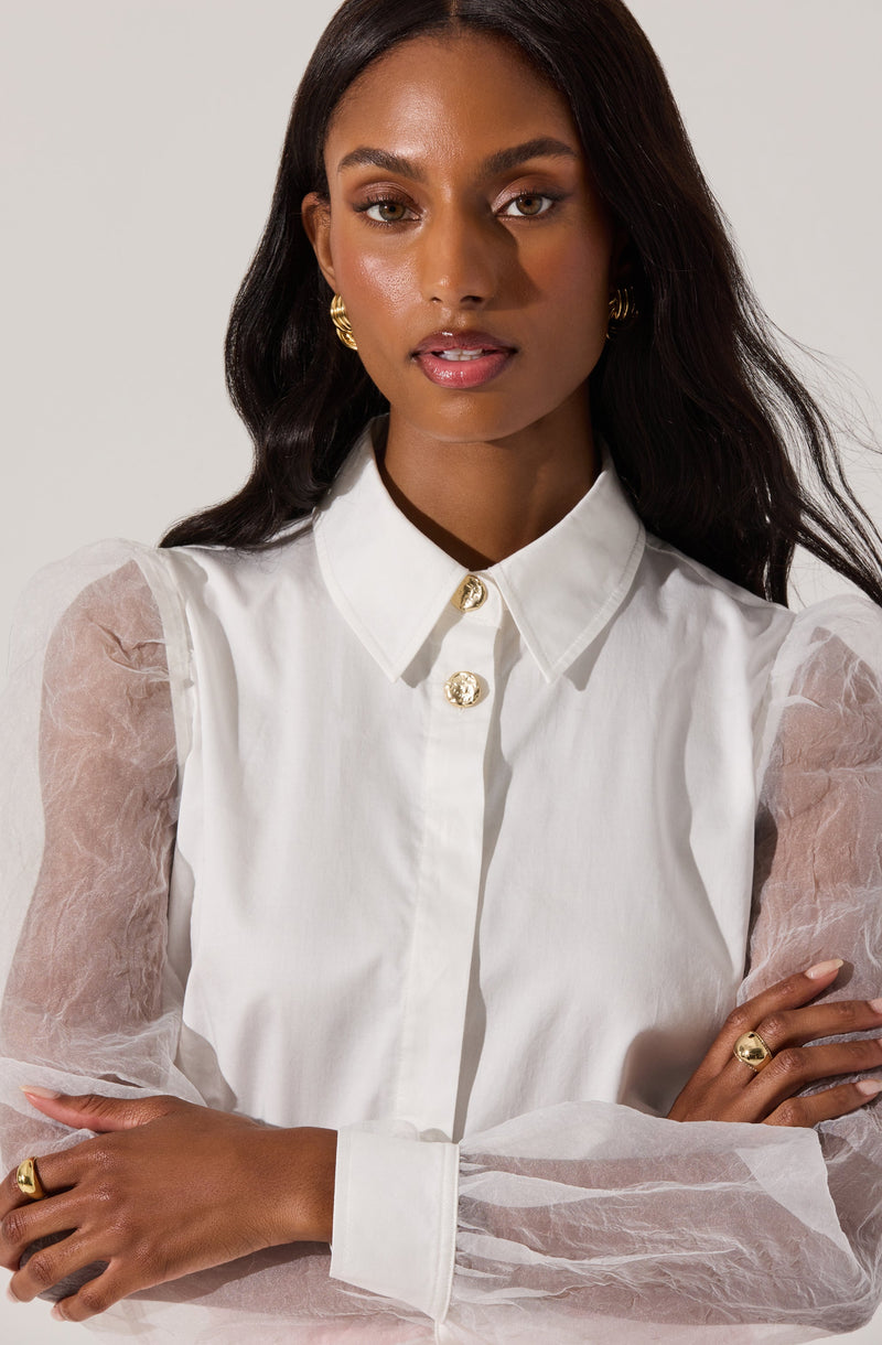 A woman wearing a white button-up shirt with a pointed collar and three decorative gold buttons at the top. The shirt has sheer, puffy long sleeves made of a translucent, slightly crinkled fabric. She has gold hoop earrings, a gold ring on her left hand, and a gold ring on her right hand.