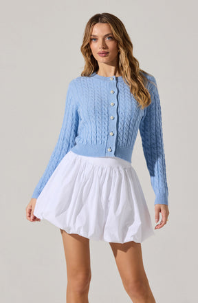 A woman is wearing a light blue, cable-knit button-up cardigan with long sleeves and a round neckline. She pairs it with a white, puffy, knee-length skirt with gathered fabric that creates a voluminous, textured appearance.