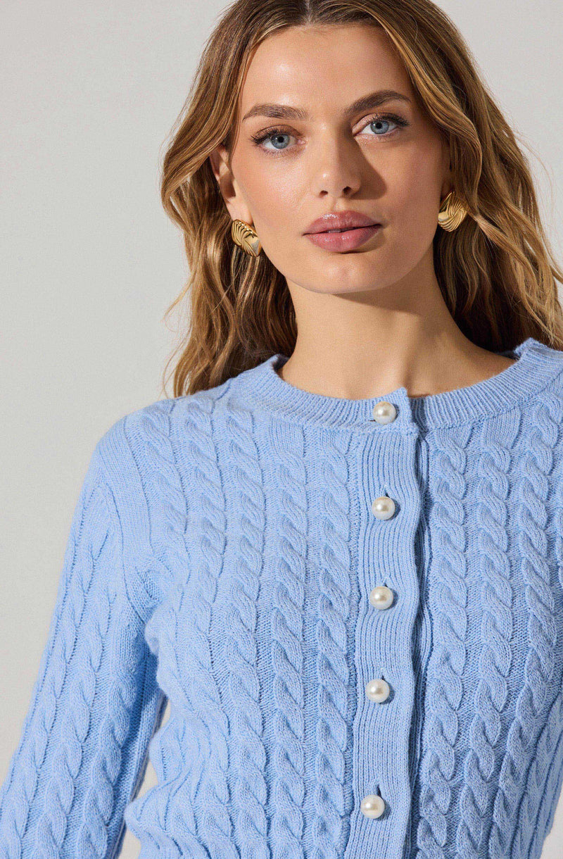 A woman with wavy, light brown hair is wearing a light blue, cable-knit sweater with a round neckline. The sweater has decorative white pearl buttons down the front. She is also wearing gold, textured hoop earrings.