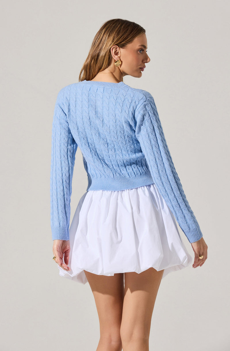 A woman is wearing a light blue, long-sleeved, cable-knit sweater with a ribbed hem and cuffs. She pairs it with a white, puffy, knee-length skirt with gathered fabric. She has a gold earring visible on her left ear.