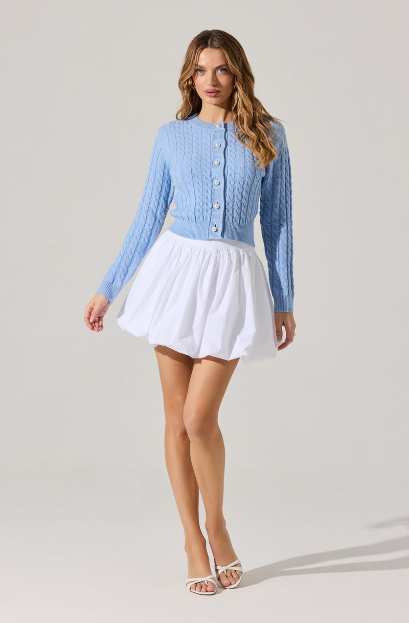 A woman is wearing a light blue, long-sleeved, cable-knit cardigan with a round neckline and buttons down the front, paired with a short, white, pleated skirt. She is also wearing white high-heeled sandals.