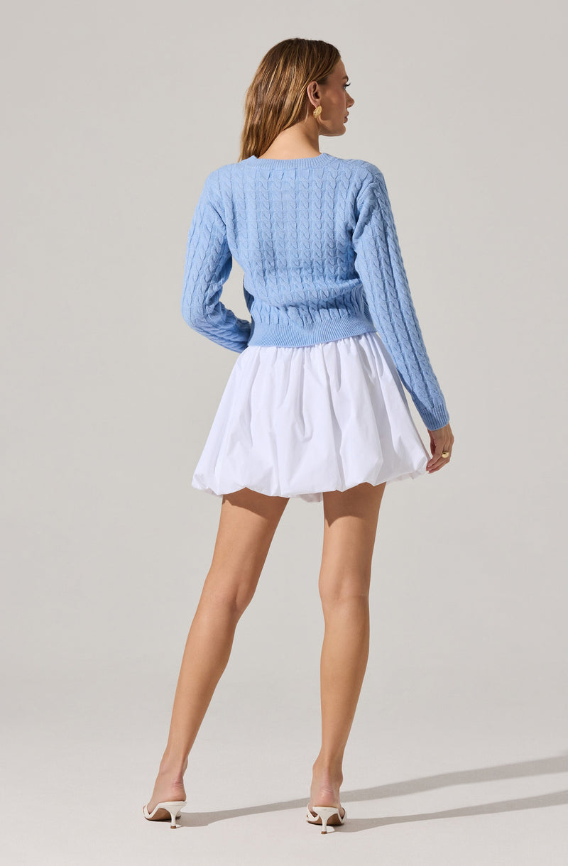 A woman is wearing a light blue cable-knit sweater with a fitted waistband and long sleeves. She is also wearing a white, voluminous, pleated skirt that falls above the knees. She has on white high-heeled shoes and gold hoop earrings.