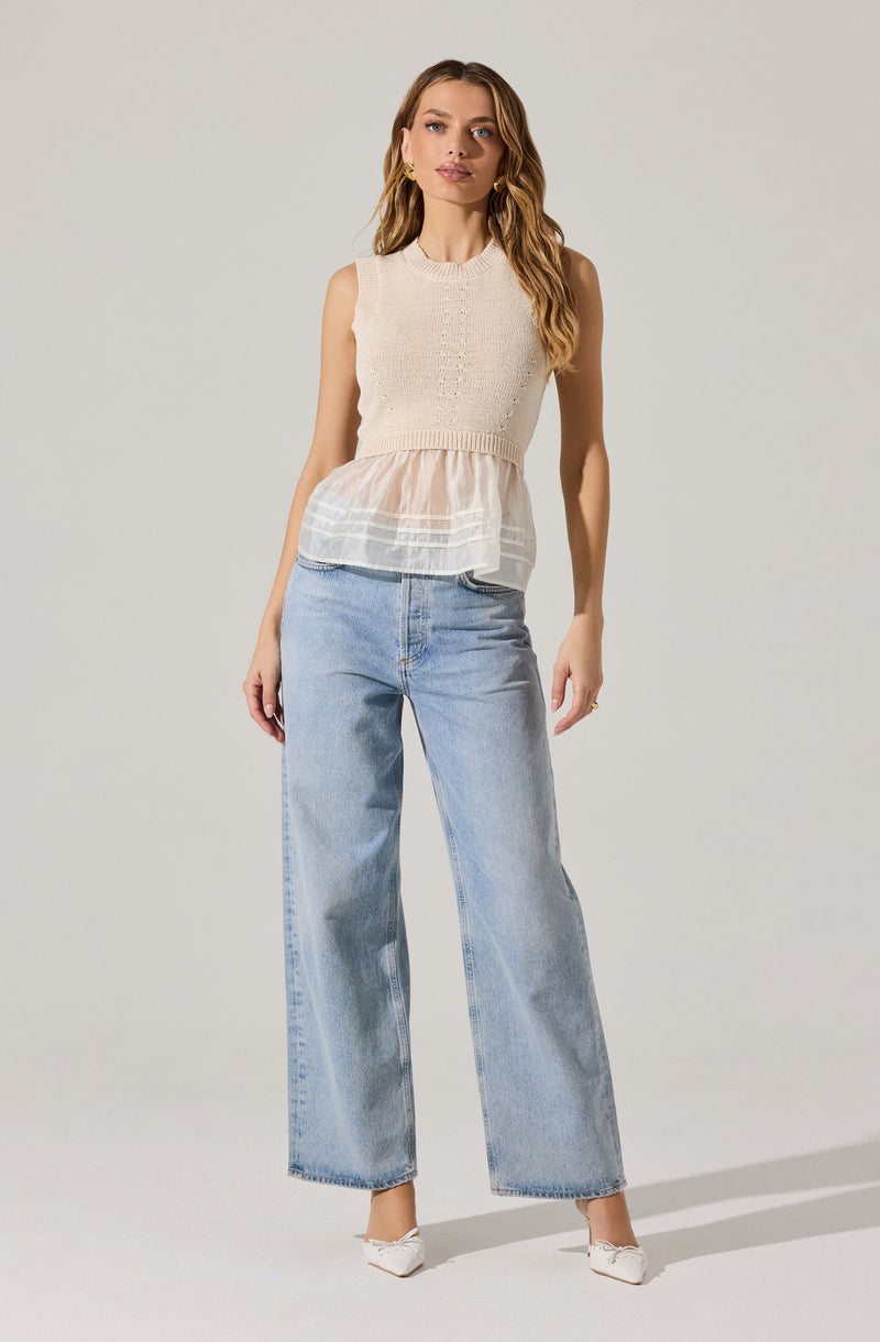 A woman is wearing a sleeveless, knitted, cream-colored top with a layered, sheer, peach-colored fabric detail at the waist. She pairs it with high-waisted, light-wash, wide-leg jeans and white pointed-toe shoes.