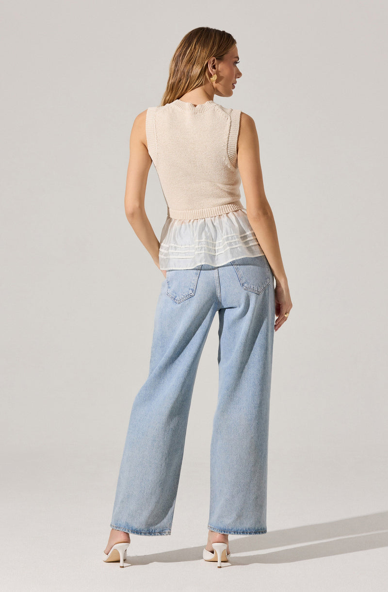 A woman is wearing a sleeveless, light-colored knit vest with a slightly cropped fit, paired with a white, sheer, ruffled top underneath. She is also wearing high-waisted, light-wash wide-leg jeans and white high-heeled shoes.