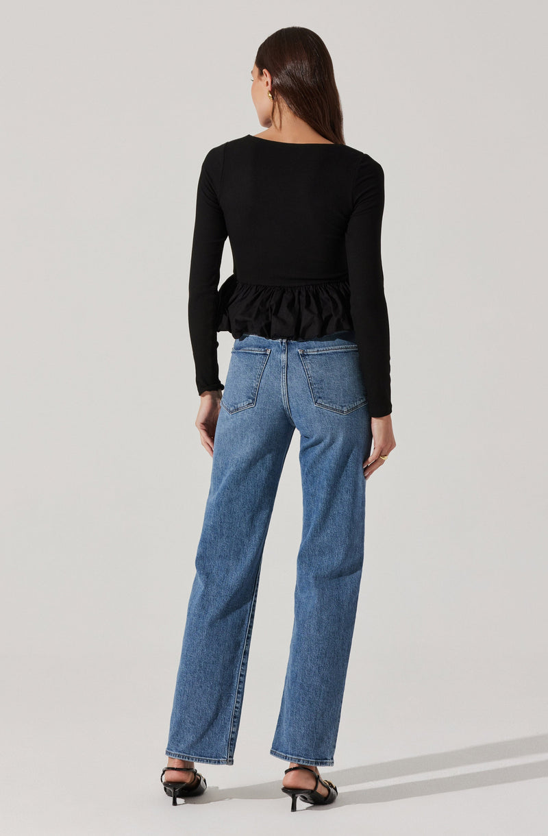 A woman is wearing a black long-sleeve top with a ruffled hem at the waist, paired with high-waisted, straight-leg blue jeans. She is also wearing black open-toe sandals with a small heel.