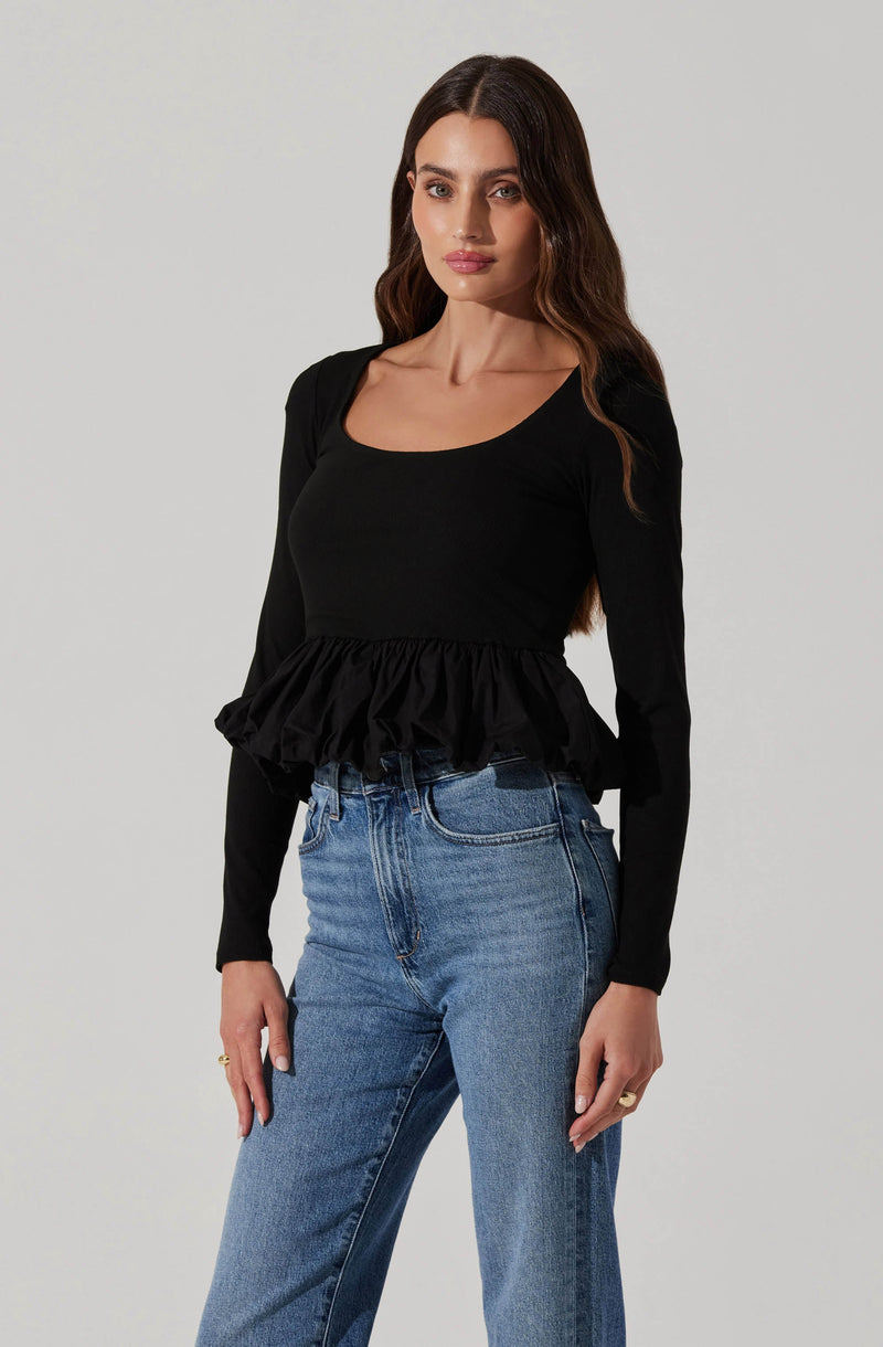 A woman is wearing a black long-sleeve top with a wide scoop neckline and a gathered ruffle detail at the hem. The top fits snugly around her torso. She is also wearing high-waisted blue jeans with a classic five-pocket design and a slightly faded wash.