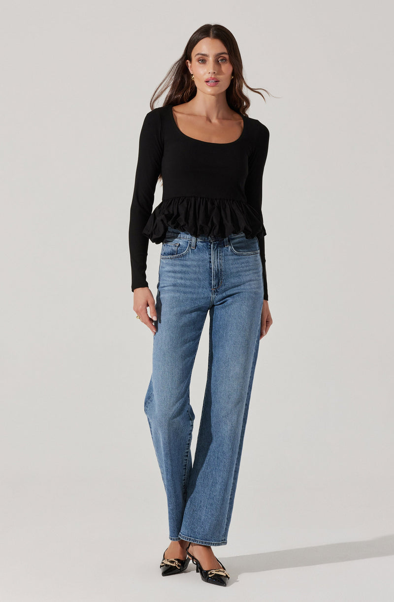 A woman is wearing a black long-sleeved top with a scoop neckline and a ruffled peplum hem. She is also wearing high-waisted, straight-leg blue jeans. Her shoes are black with a bow detail on the front and a small heel.