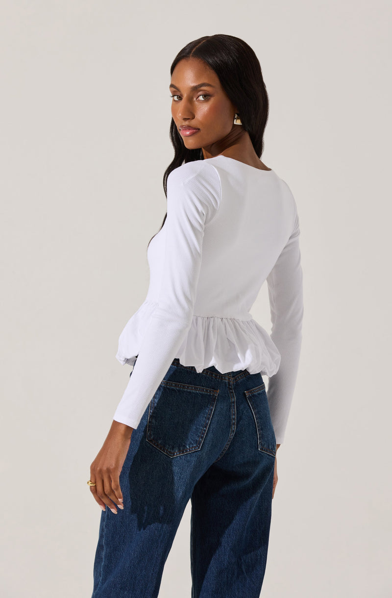 A woman is wearing a white long-sleeve top with a ruffled hem at the waist. The top has a fitted style, with the ruffles adding a decorative touch. She pairs it with high-waisted dark blue jeans.