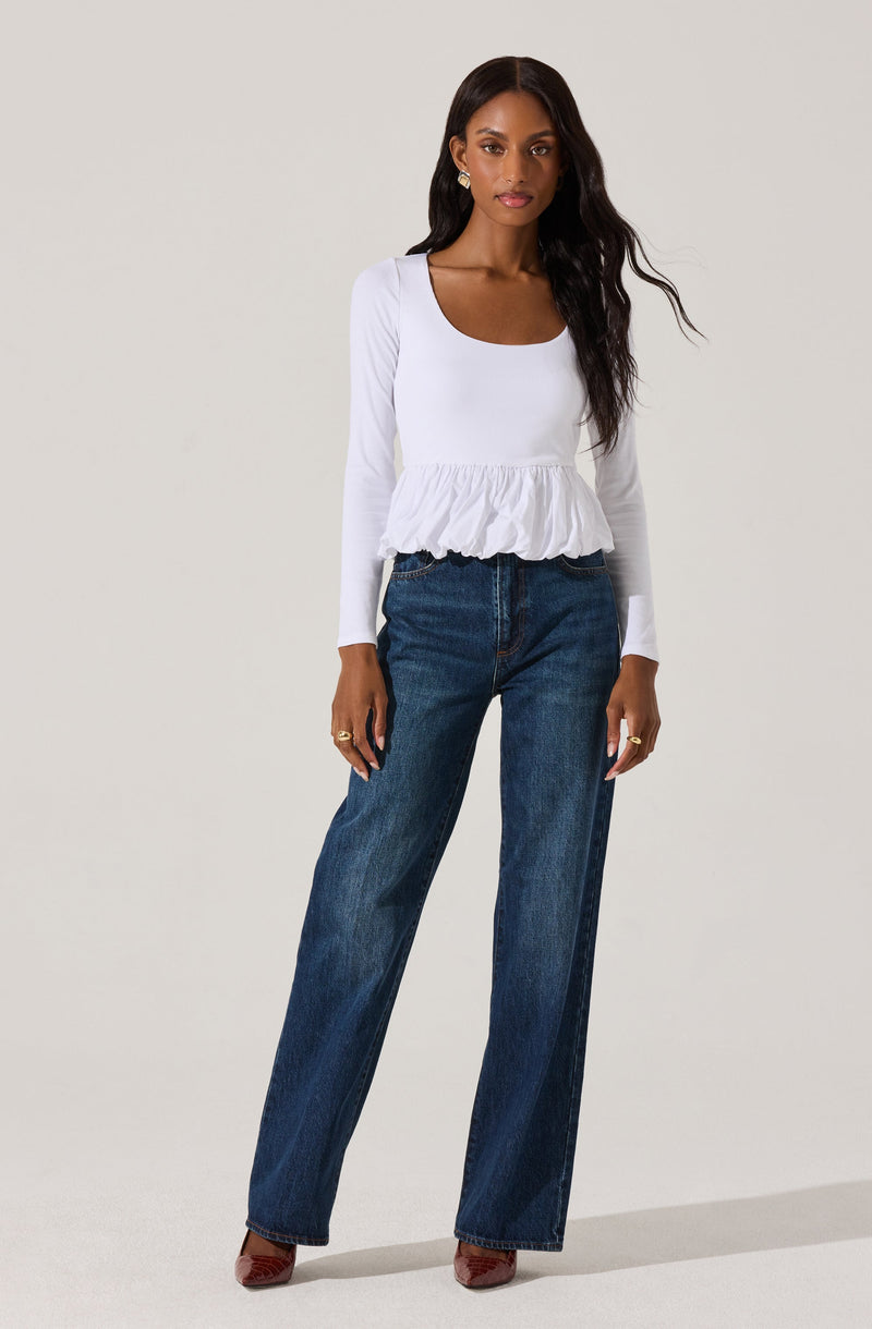 A woman is wearing a white long-sleeve top with a scoop neckline and a ruffled peplum hem. She is paired it with high-waisted, dark wash wide-leg jeans and brown pointed-toe shoes.